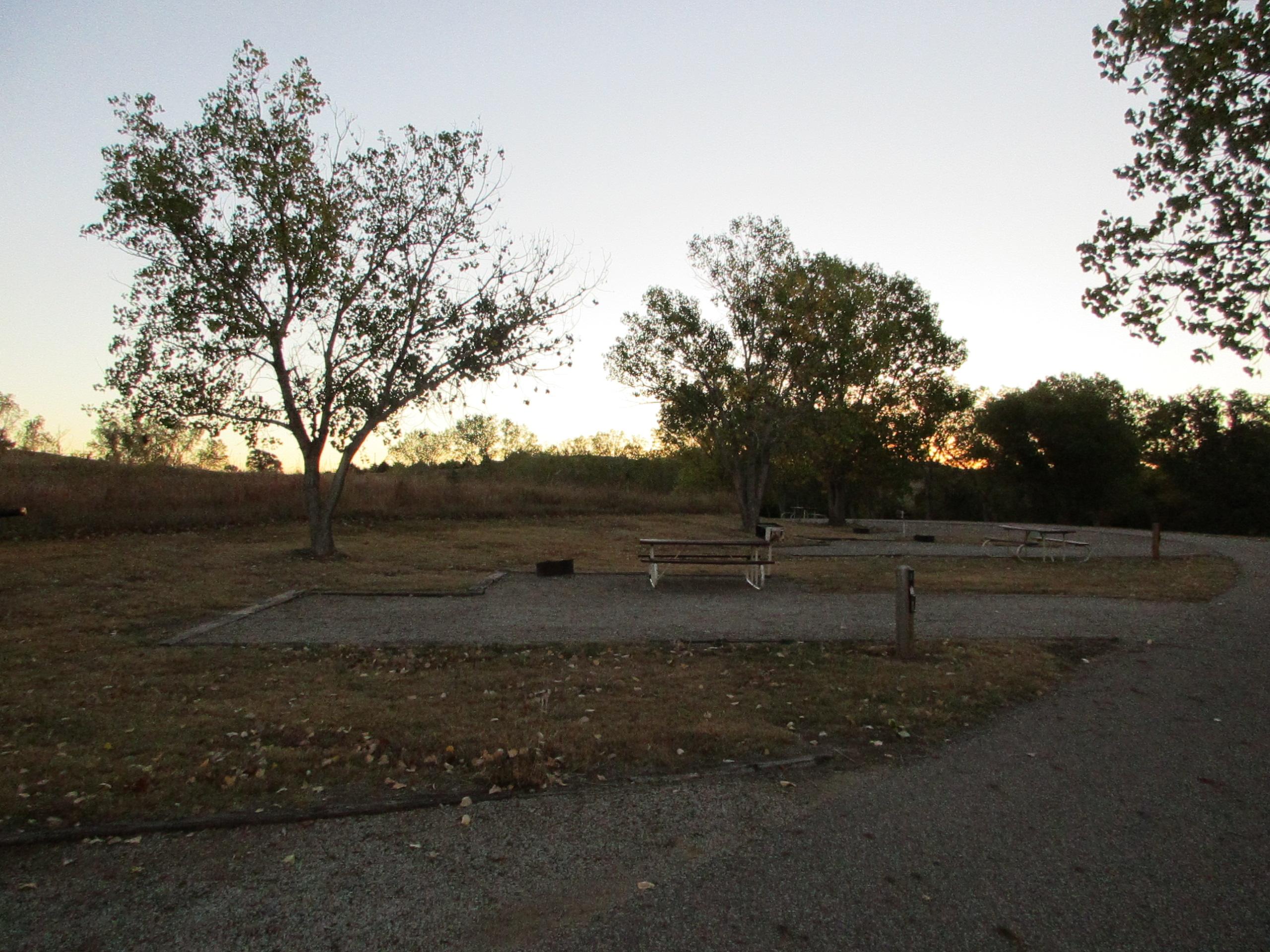 A serene outdoor scene at dusk, featuring a gravel area with several picnic tables under trees. The sky shows soft hues as the sun sets, while fallen leaves cover the ground, suggesting a tranquil and natural environment. Switchgrass mountain bike trail.