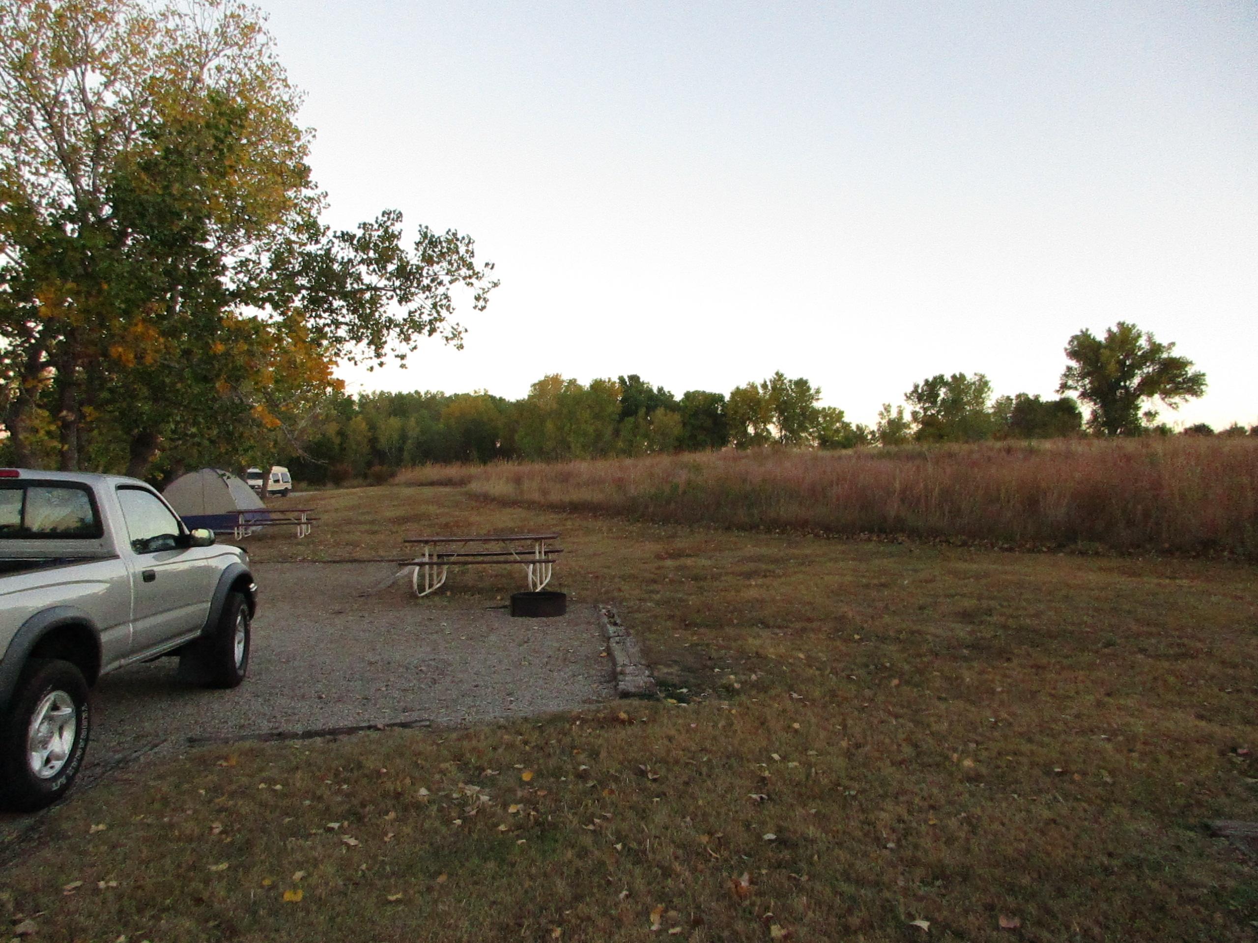 A peaceful camping site at dusk, featuring a gravel area with a parked silver pickup truck, a camping tent, and picnic tables situated on a grassy field surrounded by trees. The background shows a stretch of tall grass and additional camping areas in the distance under a soft, twilight sky. Switchgrass mountain bike trail.