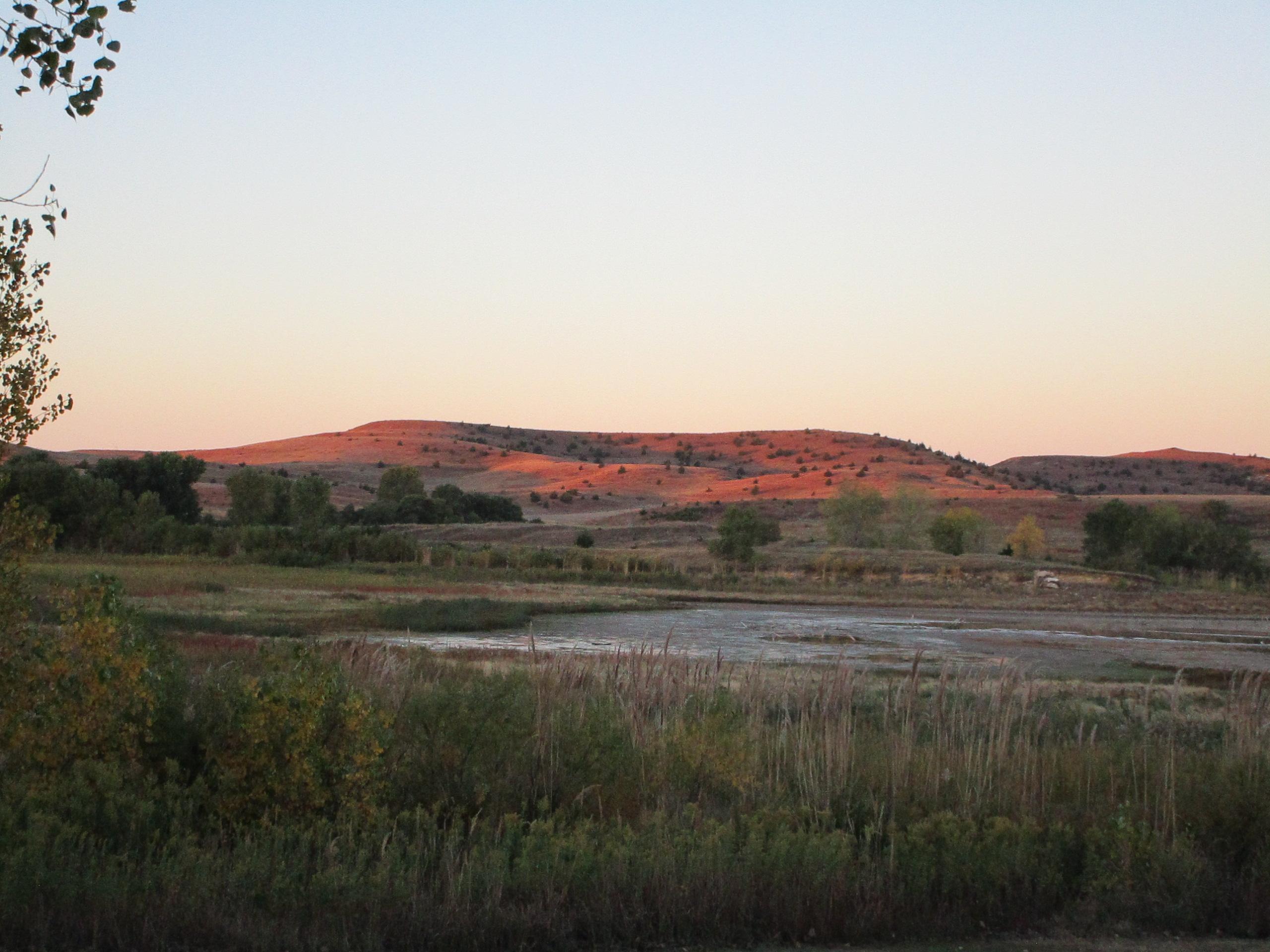 A tranquil landscape featuring rolling hills bathed in the warm glow of sunset, with a clear sky fading from blue to pale orange. In the foreground, lush greenery and tall grasses lead to a calm body of water, reflecting the serene atmosphere of the early evening. Switchgrass mountain bike trail.