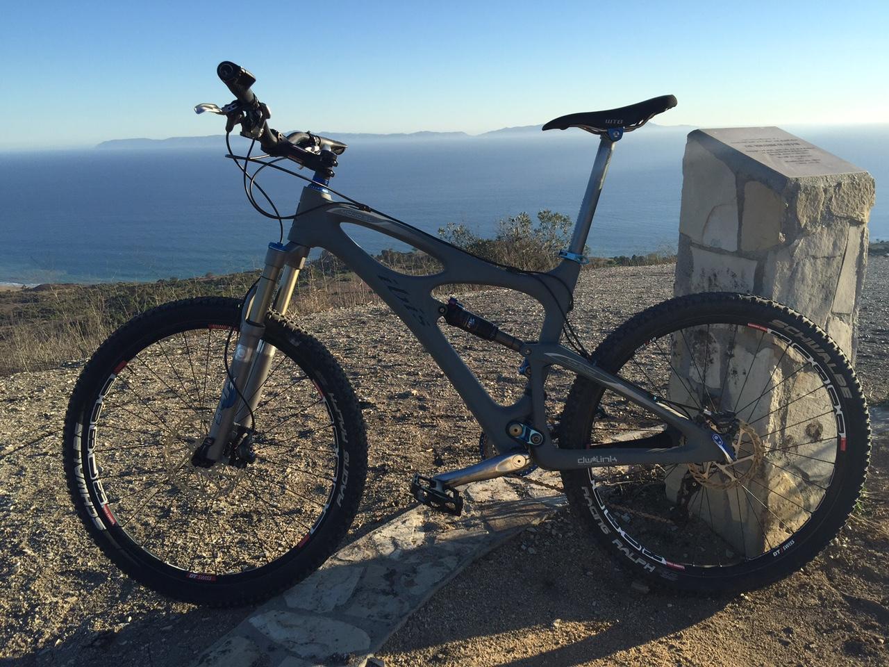 Ibis Mojo HD: A mountain bike standing on a gravel path near the edge of a cliff, overlooking the ocean. The bike has a gray frame and features front suspension. In the background, a clear blue sky meets the water, with distant land visible on the horizon. A stone pillar is nearby, adding to the scenic landscape.