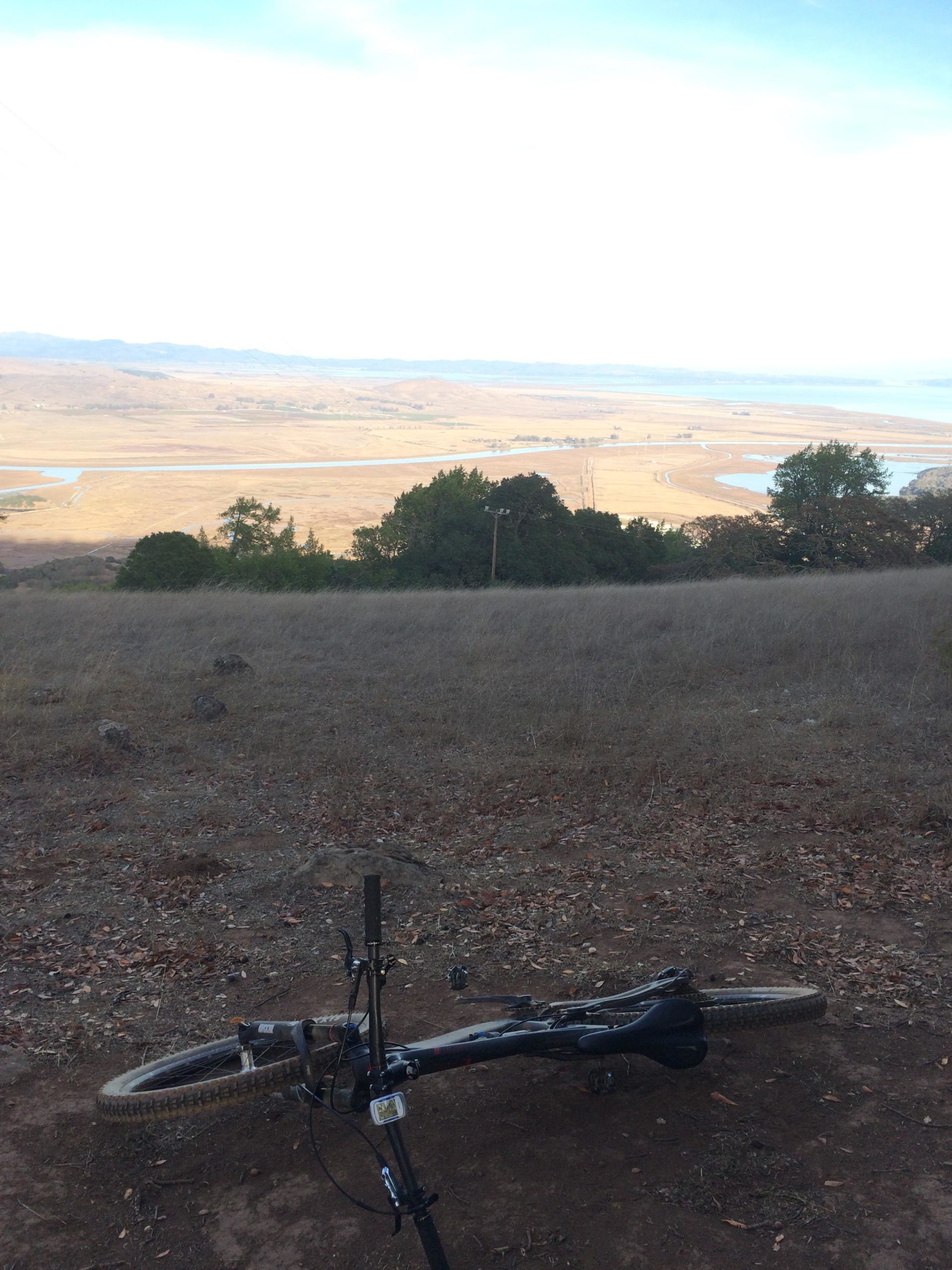 A mountain bike lying on the ground in a brown, grassy area with a scenic view of rolling hills and a body of water in the background under a partially cloudy sky. Mt Burdell mountain bike trail.