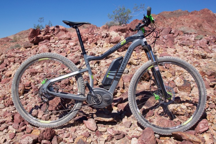 A gray and black electric mountain bike is positioned on a rocky terrain with red and brown rocks under a clear blue sky. The bike features a visible battery pack and detailed components, including a front suspension fork and wide tires designed for off-road use.