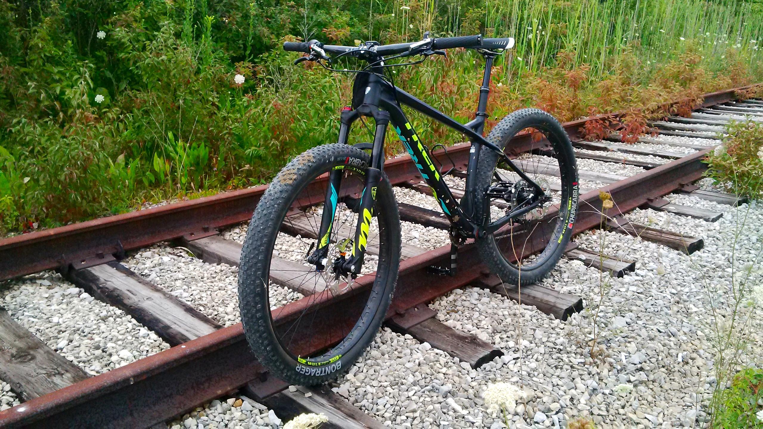 Trek Stache 9: A black and colorful mountain bike positioned on old railroad tracks, surrounded by greenery and wildflowers. The bike features thick tires and a suspension fork, resting on gravel and wooden ties.