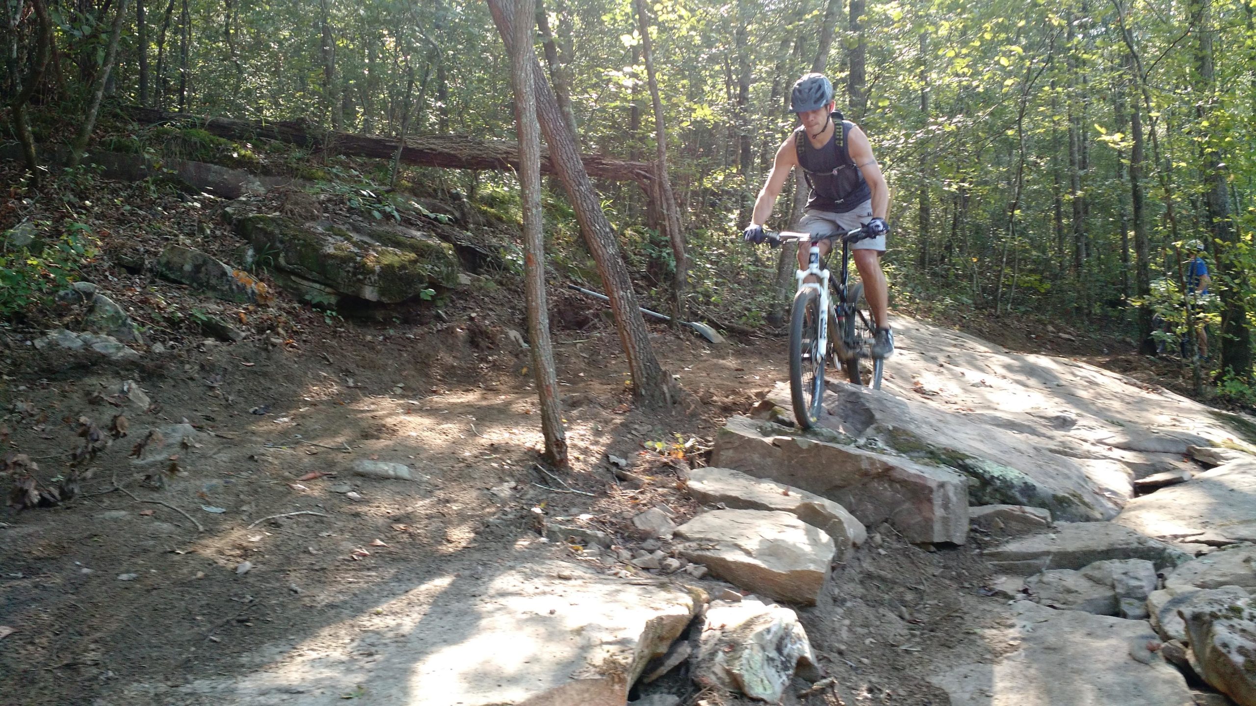 A person riding a mountain bike on a rocky trail in a forested area, surrounded by trees and sunlight filtering through the leaves. The trail features uneven terrain with large rocks and dirt. Duck River mountain bike trail.
