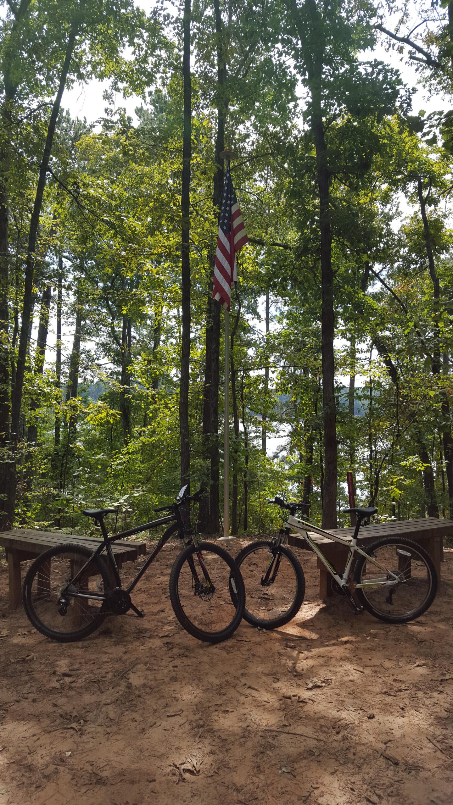 Two mountain bikes are parked on a sandy area surrounded by tall trees. In the background, an American flag is visible, gently swaying in the breeze, with a glimpse of a lake beyond the trees. The scene suggests a peaceful outdoor setting, ideal for cycling and enjoying nature. Blankets Creek mountain bike trail.