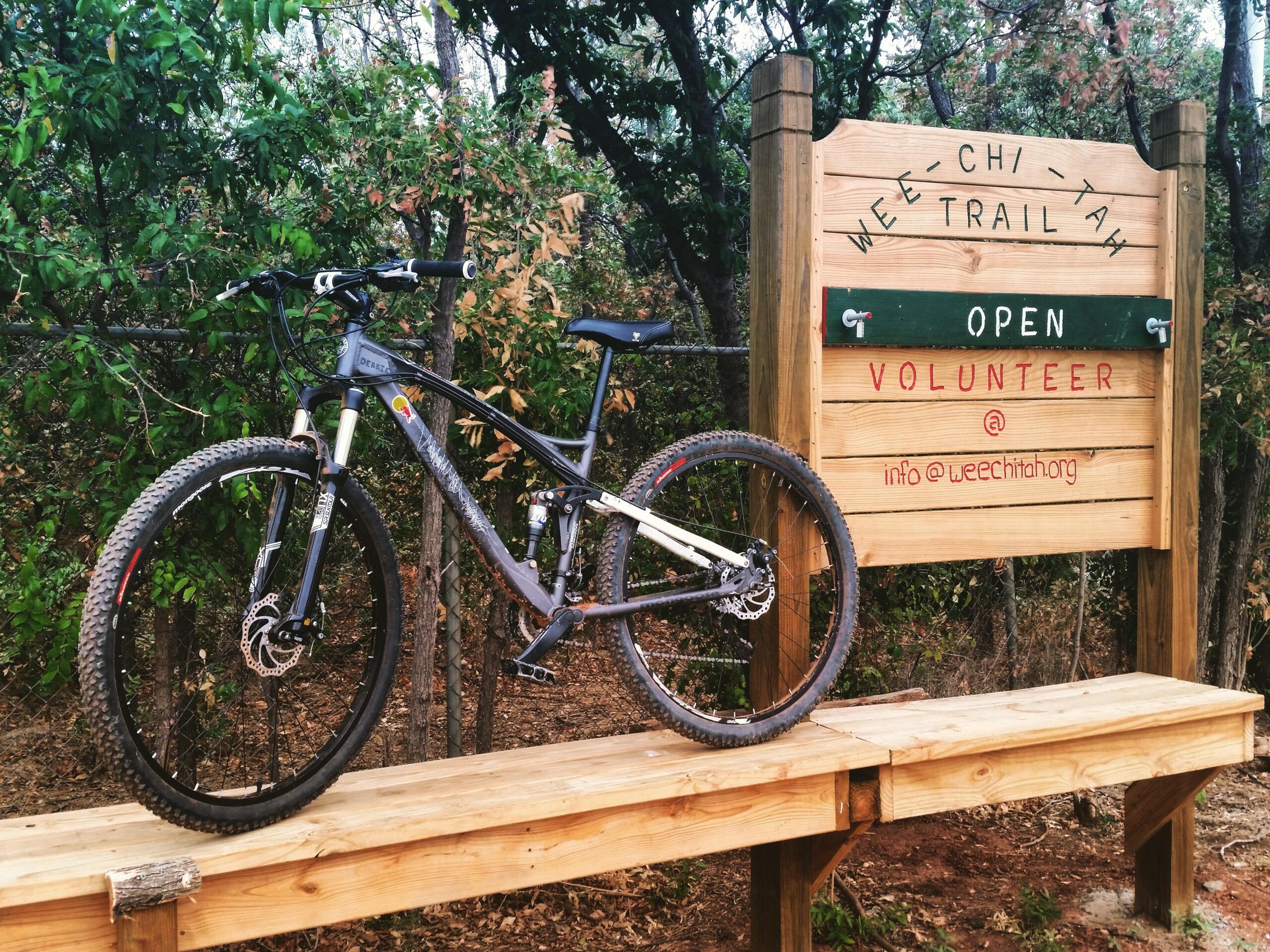 Mongoose MONGOOSE XR-PRO: Mountain bike resting on a wooden bench in front of a sign for the Wee-Chi-Tah Trail, which is open for use and encourages volunteer participation. The sign features contact information and is surrounded by lush greenery.