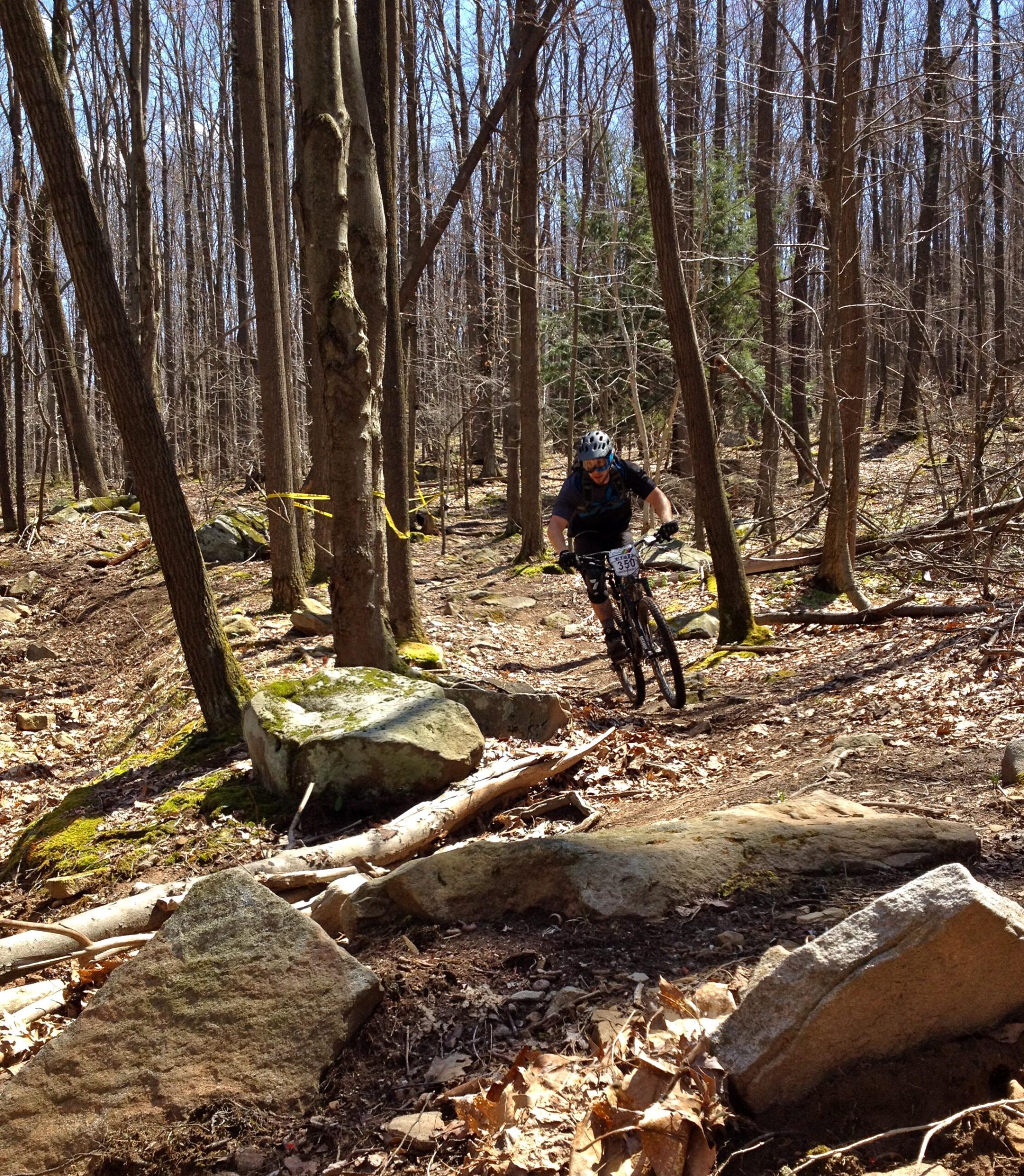 A mountain biker navigates a rocky trail through a forest of bare trees on a sunny day. The terrain features large stones, fallen branches, and patches of leaves scattered on the ground. Yellow tape can be seen marking the trail. Big Bear Lake Trail Center mountain bike trail.