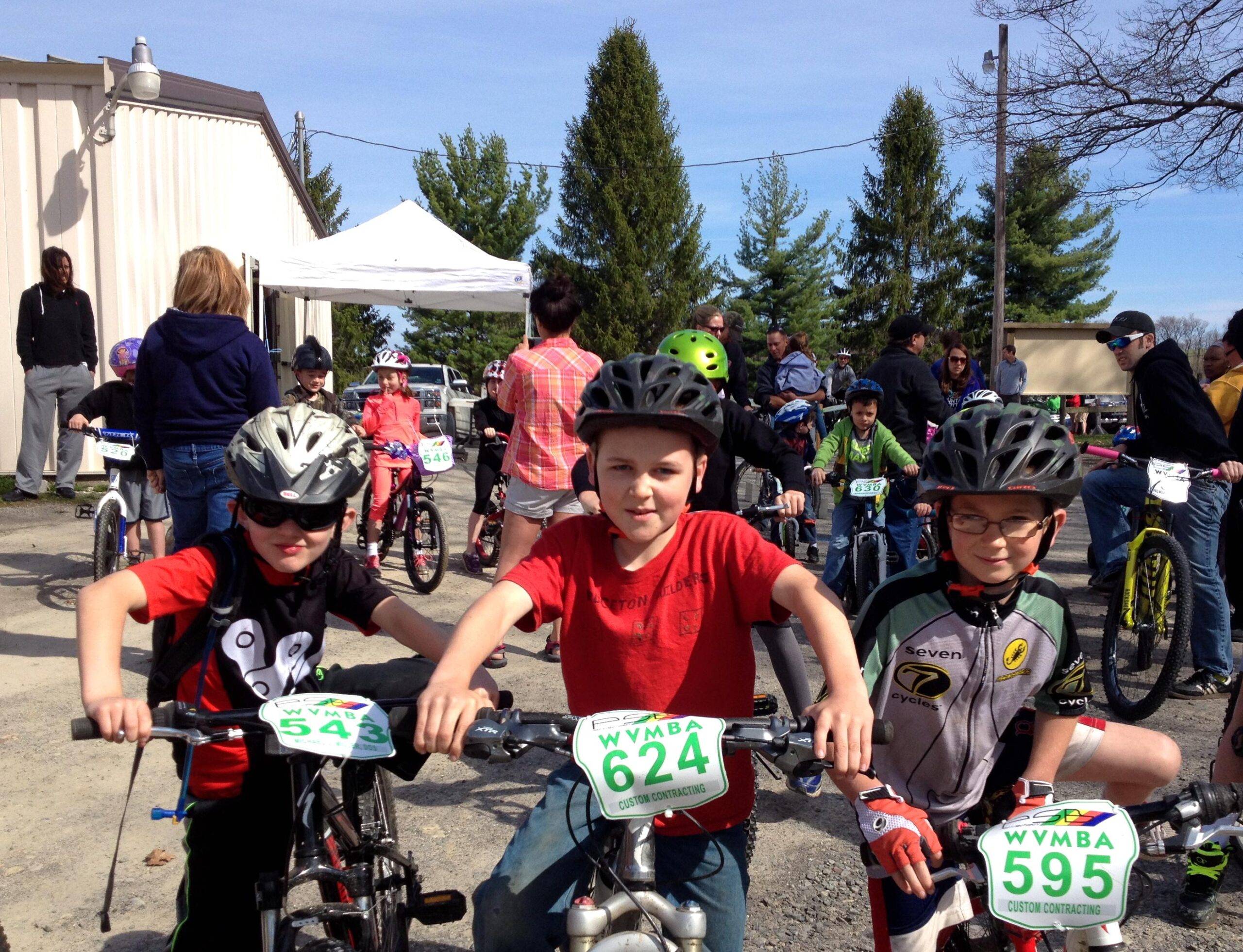 A group of three children wearing helmets and biking attire pose with their bicycles at a cycling event. The child on the left is wearing a red shirt with a bear graphic, the middle child has a red shirt, and the child on the right is in a black and white cycling jersey. In the background, other kids are preparing for the race, with a tent and spectators visible. The scene is set outdoors on a sunny day. Big Bear Lake Trail Center mountain bike trail.
