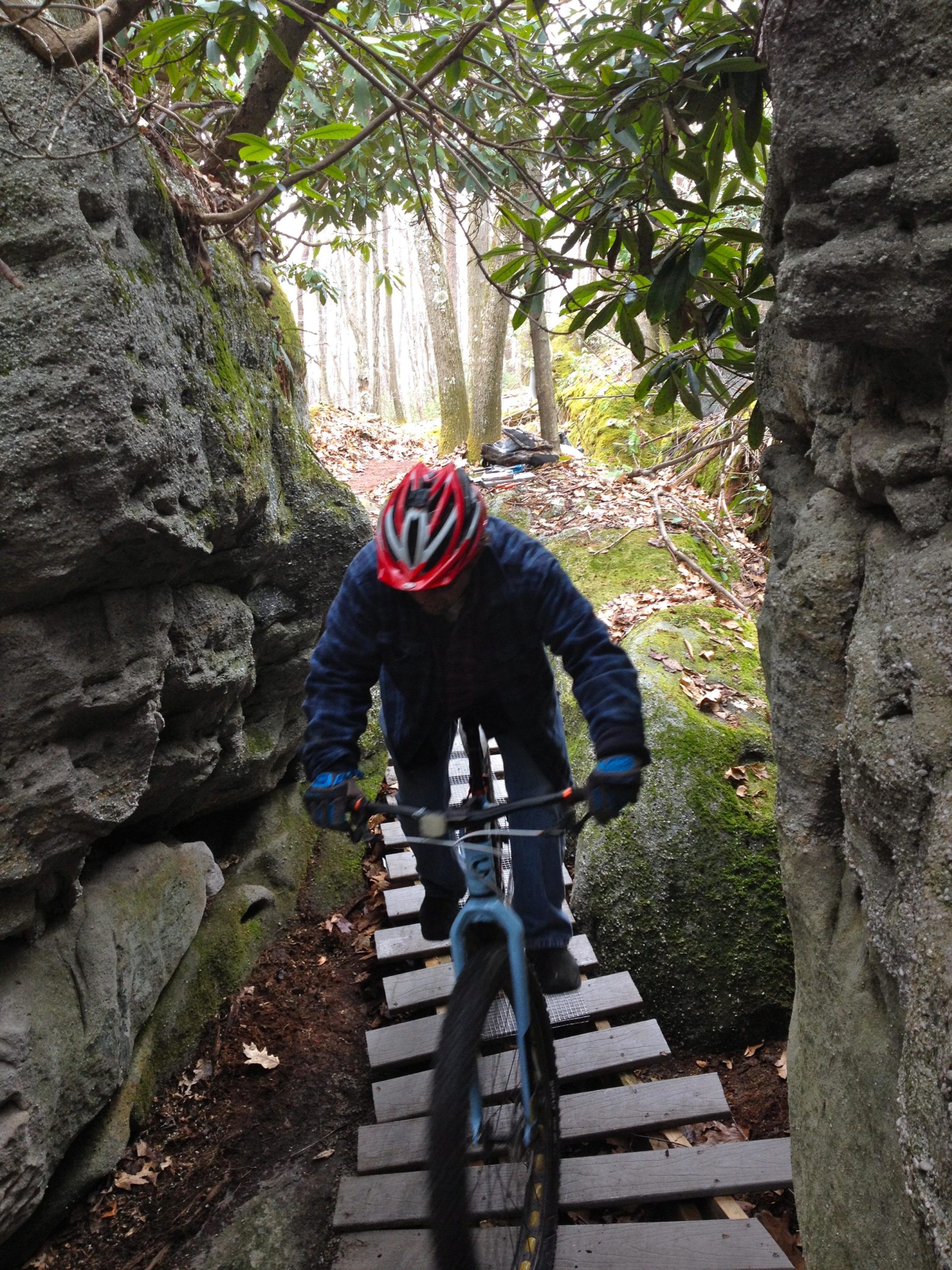 A mountain biker traverses a narrow wooden bridge between two large rocks in a forested area, surrounded by trees and greenery. The rider is wearing a red helmet and a blue jacket as they navigate the rugged terrain. Big Bear Lake Trail Center mountain bike trail.