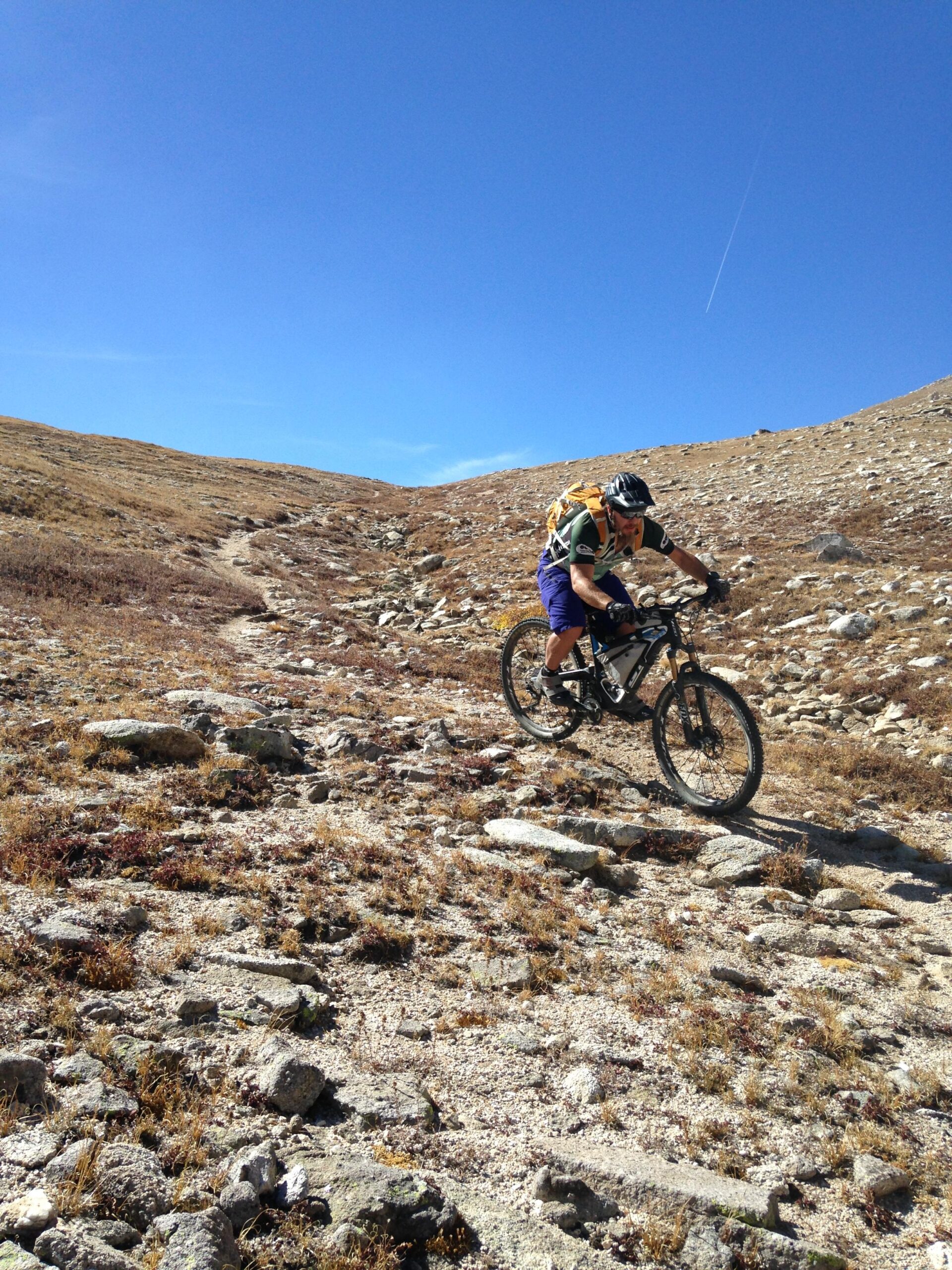 A mountain biker rides over a rocky trail in a mountainous terrain under a clear blue sky. The landscape is mostly dry with patches of grass, and the biker is wearing a helmet and a backpack, focused on navigating the challenging path. Little Browns Creek mountain bike trail.