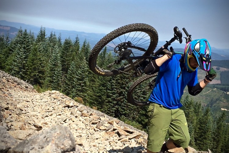 A mountain biker wearing a colorful helmet and blue shirt carries their bike up a rocky trail surrounded by tall evergreen trees, with hills visible in the distance.