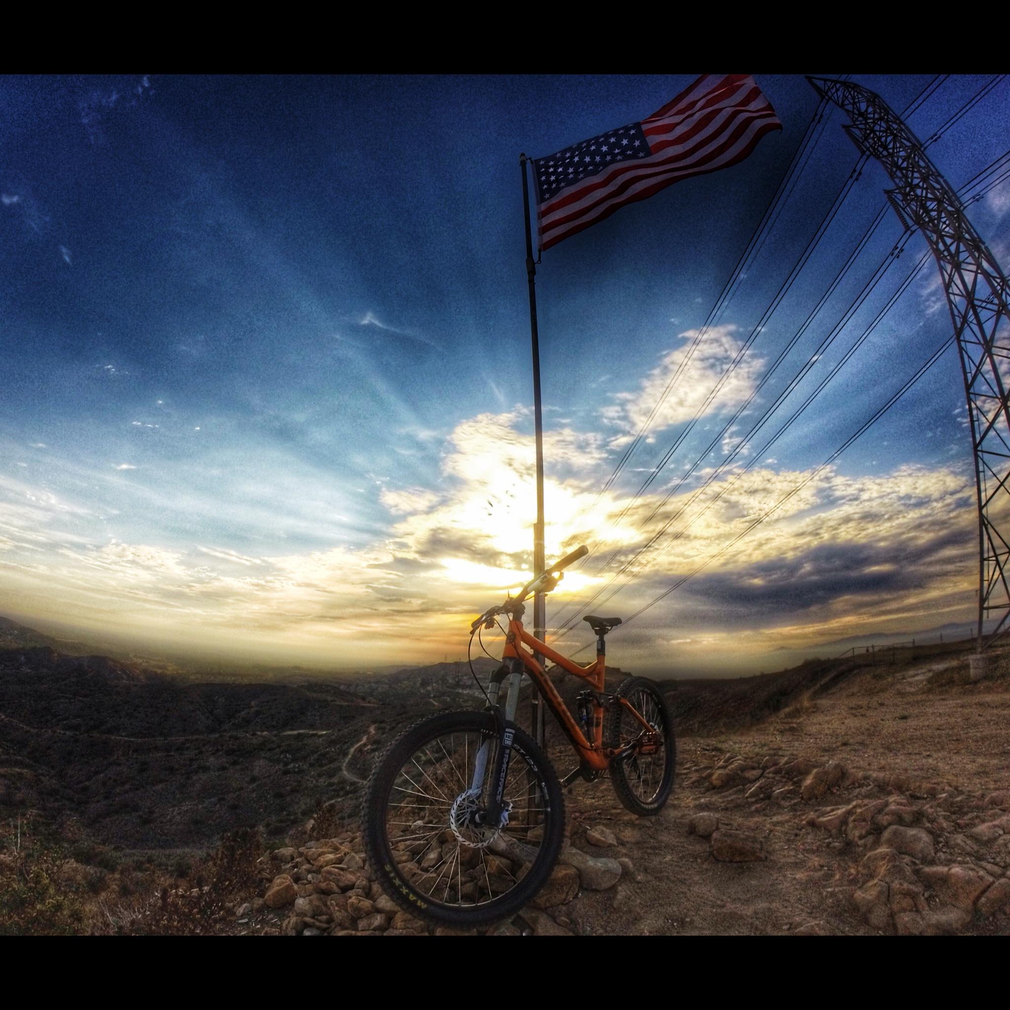 Kona Process 153: An orange mountain bike rests on rocky terrain with a backdrop of a vibrant sunset. A tall American flag flies next to the bike, while power lines stretch across the sky above. The scene captures a moment of tranquility in nature, highlighting the expansive landscape and colorful clouds.
