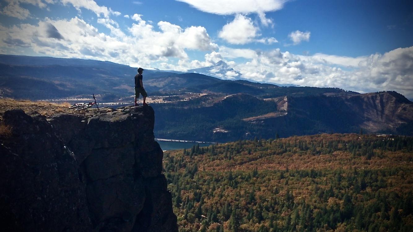 A person standing on the edge of a rocky cliff, overlooking a vast landscape of rolling hills and valleys covered in trees, with a river winding through the scene. The sky is partly cloudy, and a distant snow-capped mountain is visible on the horizon. A bicycle is resting beside the cliff. Syncline mountain bike trail.