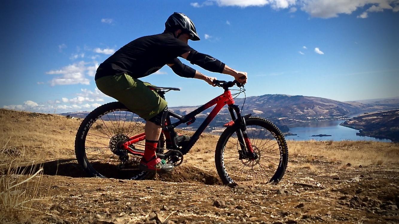 A person riding a mountain bike on a rugged terrain, leaning forward with determination. The landscape features dry grass and hills, with a body of water visible in the background under a clear blue sky filled with scattered clouds. Syncline mountain bike trail.
