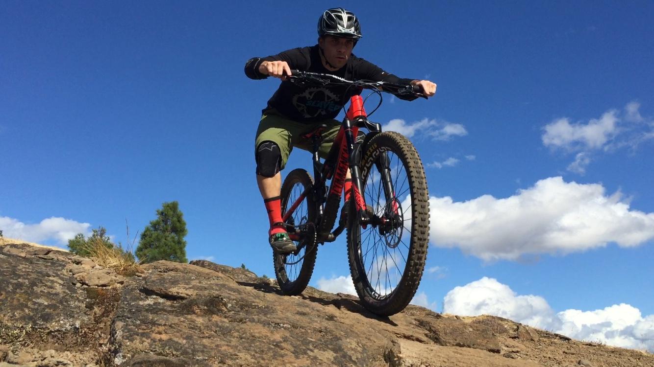 A person riding a mountain bike on rocky terrain, leaning forward with a focused expression. The background features a clear blue sky with white clouds and some greenery. The cyclist is wearing a black shirt, green shorts, and protective gear. Syncline mountain bike trail.