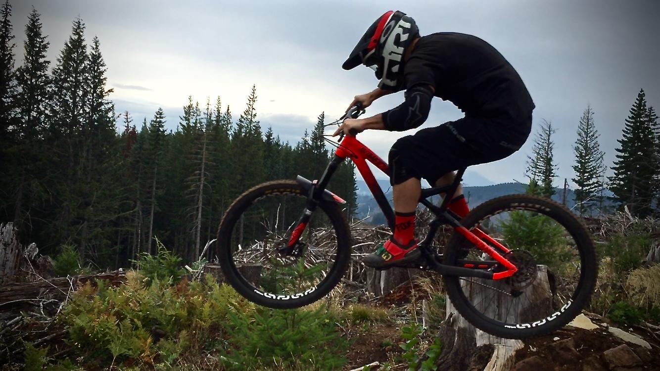 A mountain biker in a black helmet and gear performs a jump over a log in a wooded area, surrounded by tall evergreen trees and a cloudy sky. The biker's red and black bike is mid-air as they navigate an outdoor trail. Cold Creek mountain bike trail.