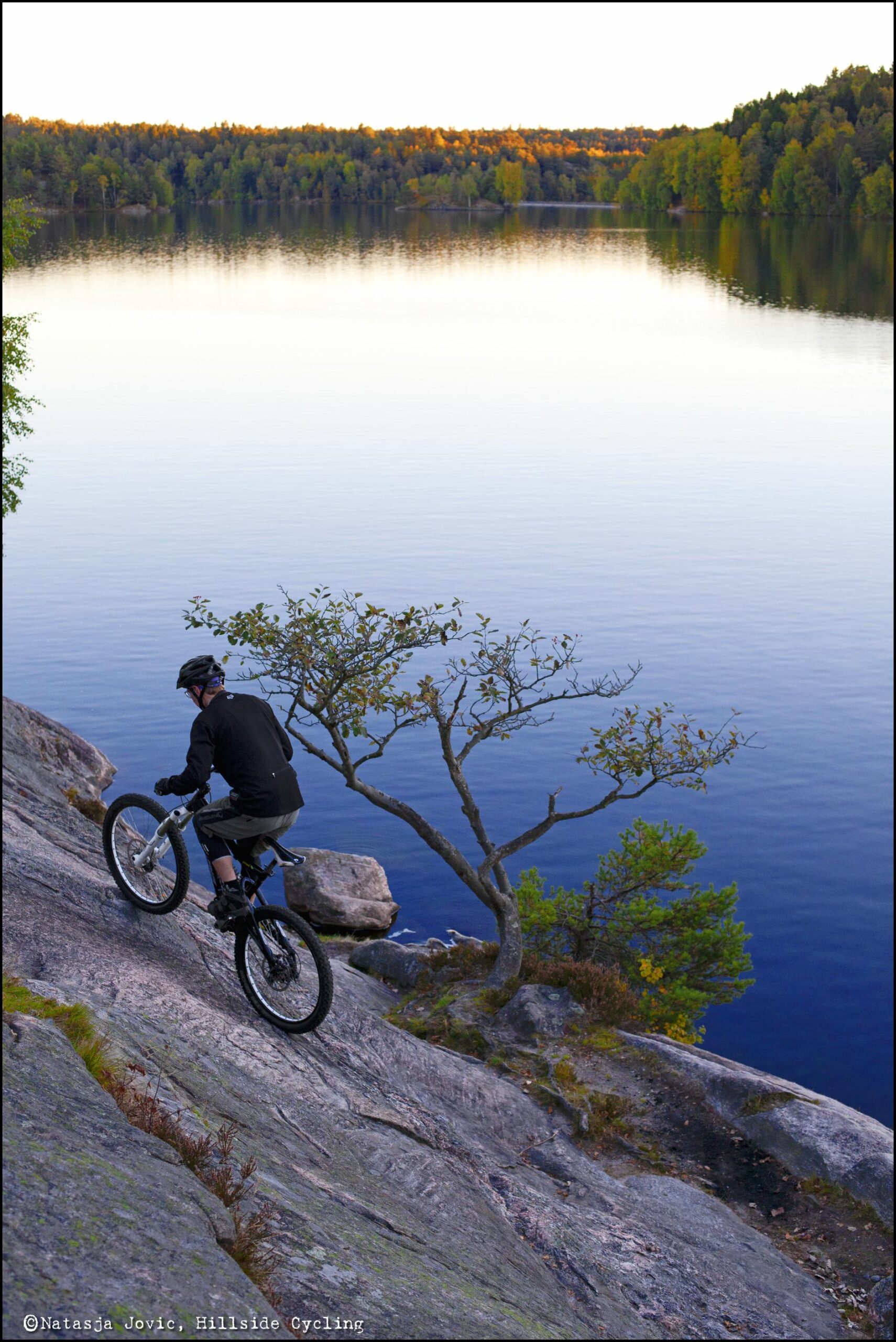 A mountain biker navigating down a rocky slope, with a calm lake and autumnal trees in the background. The scene captures the beauty of nature alongside an adventurous cycling moment. Delsjon Area mountain bike trail.