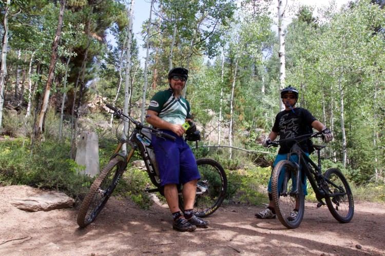 Two mountain bikers are pictured standing next to their bikes on a trail surrounded by lush greenery and aspen trees. One biker, wearing a green and white jersey, holds a small device in his hand, while the other, dressed in a black t-shirt and blue shorts, stands beside his bike with a water tube visible. The scene conveys a relaxed moment in nature, showcasing the enjoyment of outdoor biking.