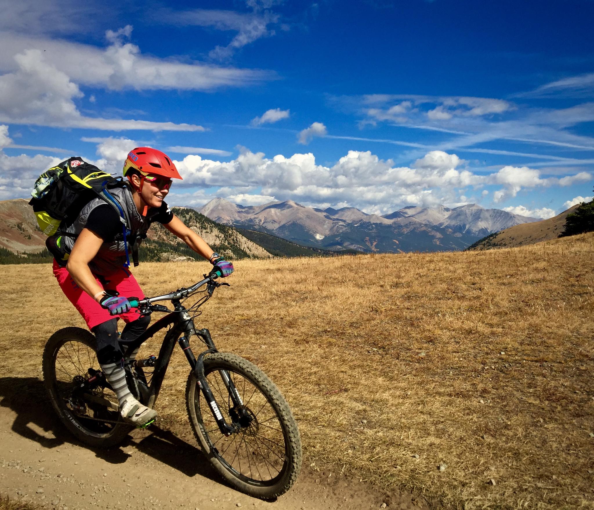 A mountain biker wearing a helmet and colorful attire rides along a dirt trail through a grassy landscape. In the background, majestic mountains rise under a blue sky with fluffy clouds. The scene conveys a sense of adventure and outdoor activity. Monarch Crest Trail mountain bike trail.