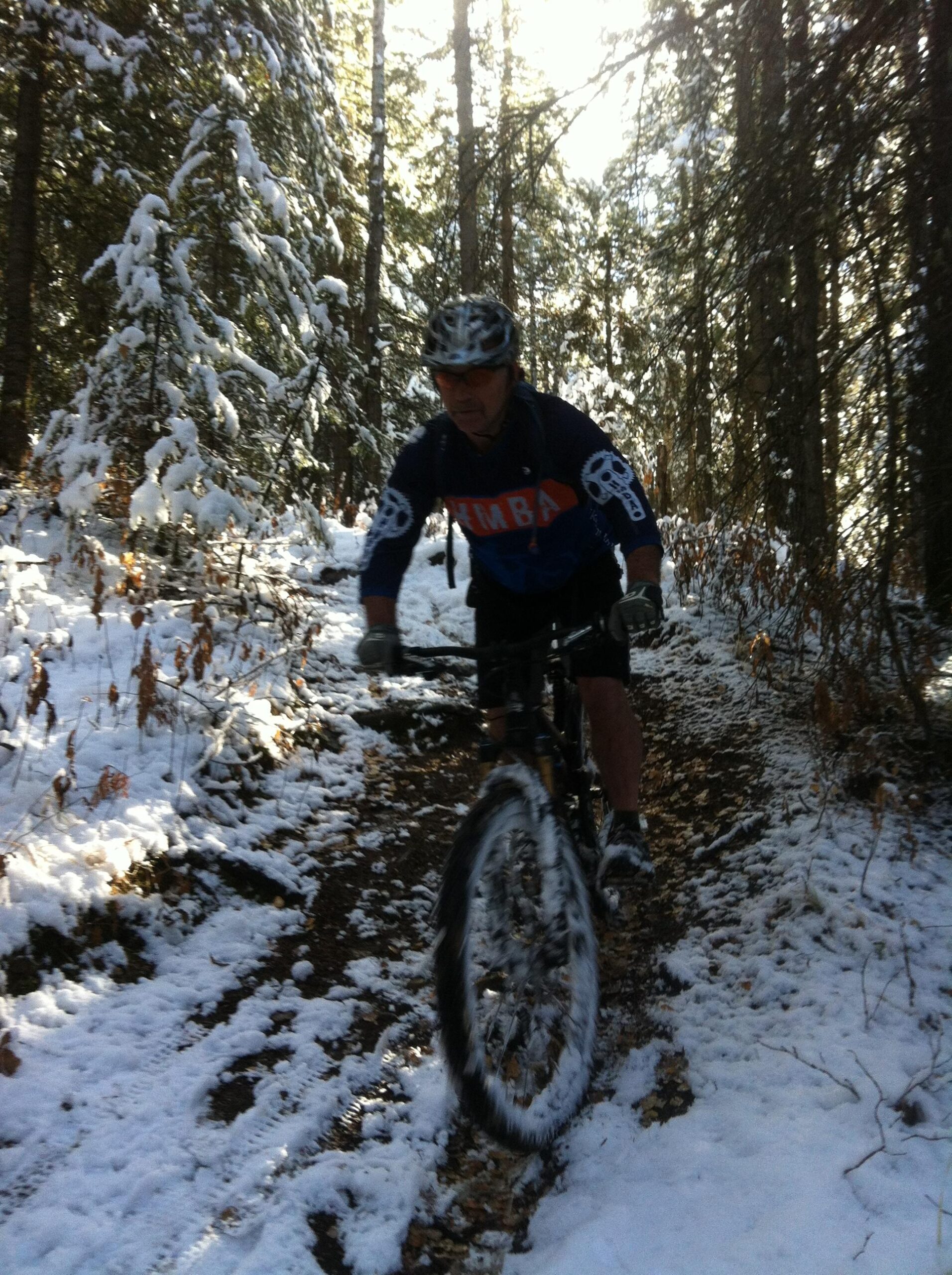 A mountain biker navigating a snowy trail in a forest. The rider wears a helmet and a long-sleeved shirt, with snow covering the ground and hanging on the trees. Sunlight filters through the trees, creating a serene winter atmosphere. Big Horn mountain bike trail.