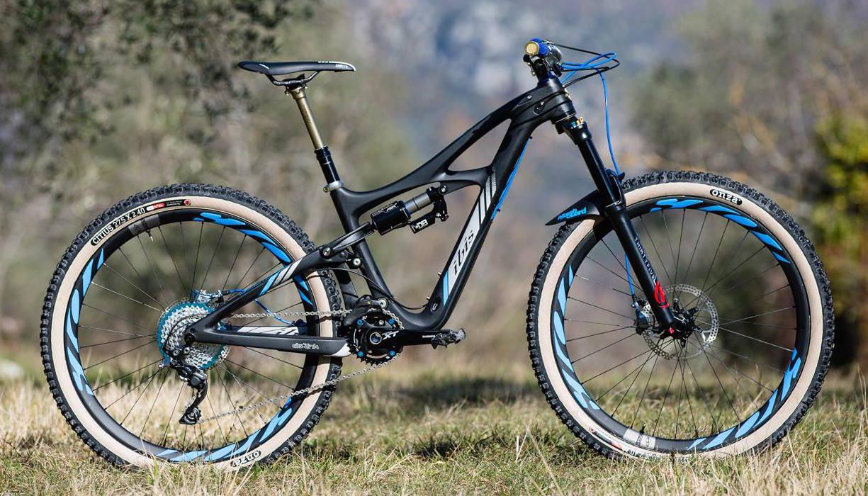 Ibis Mojo HD3: A black mountain bike with blue accents standing on grass, showcasing its sleek frame and high-performance features. The bike has knobby tires for off-road terrain and is positioned in an outdoor setting with blurred trees and a natural background.