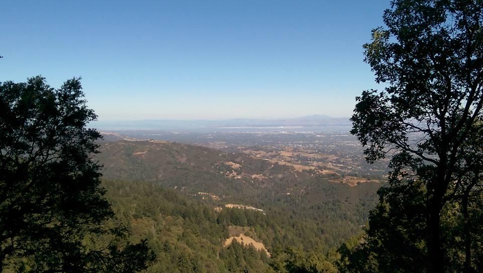 A panoramic view of rolling hills and valleys under a clear blue sky, framed by leafy trees on either side. The landscape features a mix of greenery and exposed terrain, with distant mountains visible on the horizon, creating a serene natural scene. John Nicholas mountain bike trail.