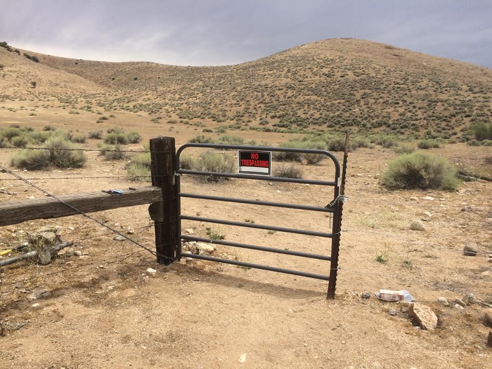 A gray metal gate with a "No Trespassing" sign stands in a dry, barren landscape. In the background, rolling hills are covered with sparse vegetation, and the ground is rocky and dusty. The sky is overcast, suggesting a potential storm. TMTA Lehigh trails mountain bike trail.