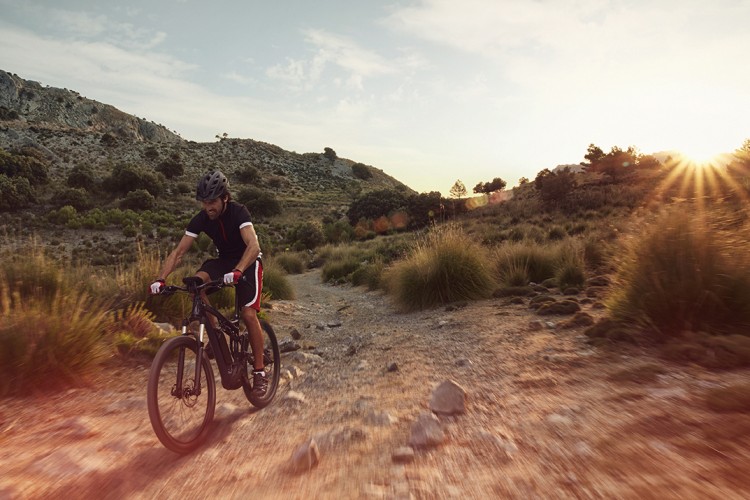 A cyclist riding a mountain bike along a rugged trail in a scenic outdoor landscape during sunset, with hills and shrubs in the background. The sunlight creates a warm glow, adding to the serene atmosphere of the setting.