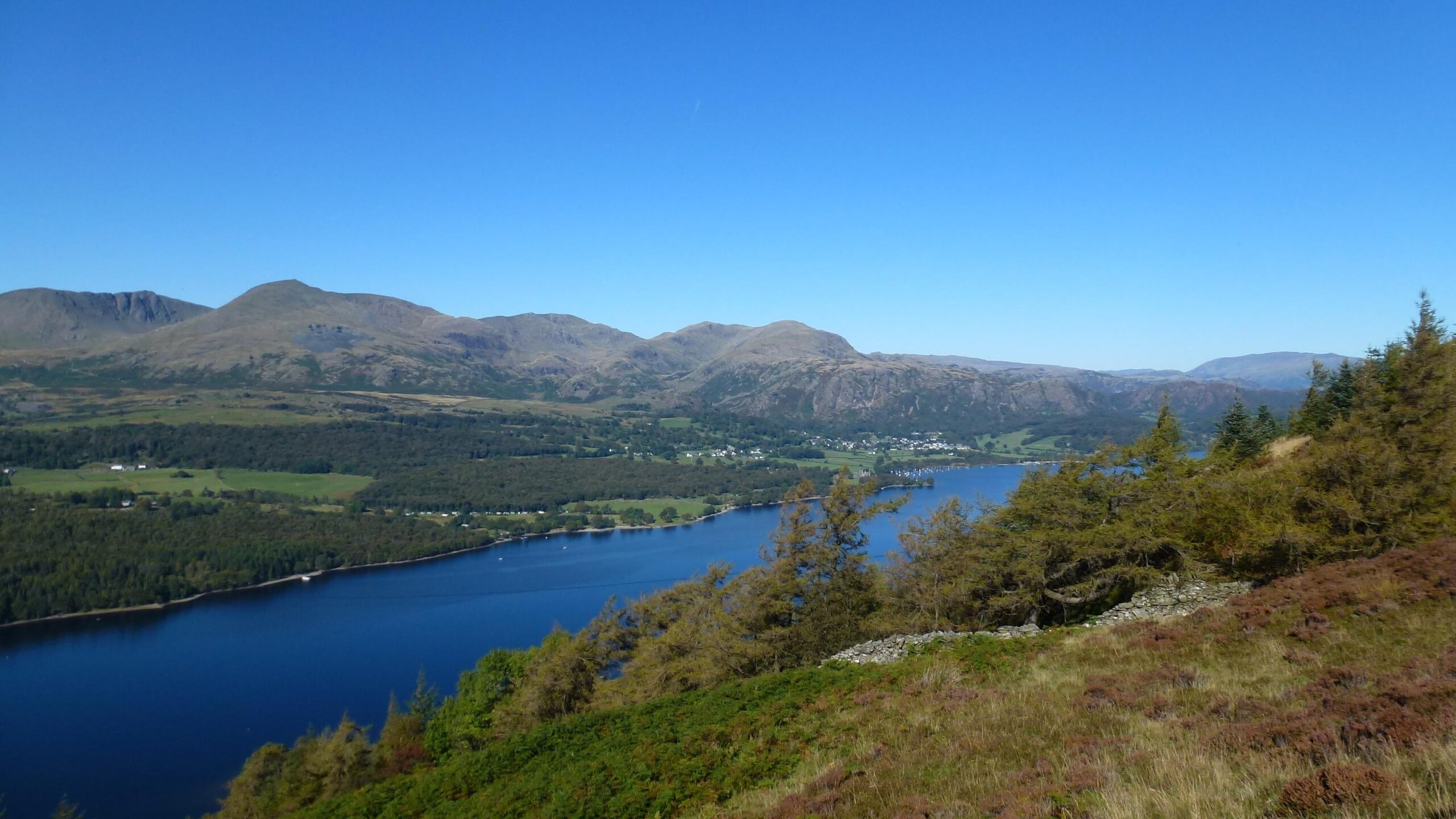 A panoramic view of a serene lake surrounded by lush green hills and mountains under a clear blue sky. The landscape features a mix of trees and grassy areas, with a small town visible along the lake's shore. Grizedale's North Face Trail mountain bike trail.