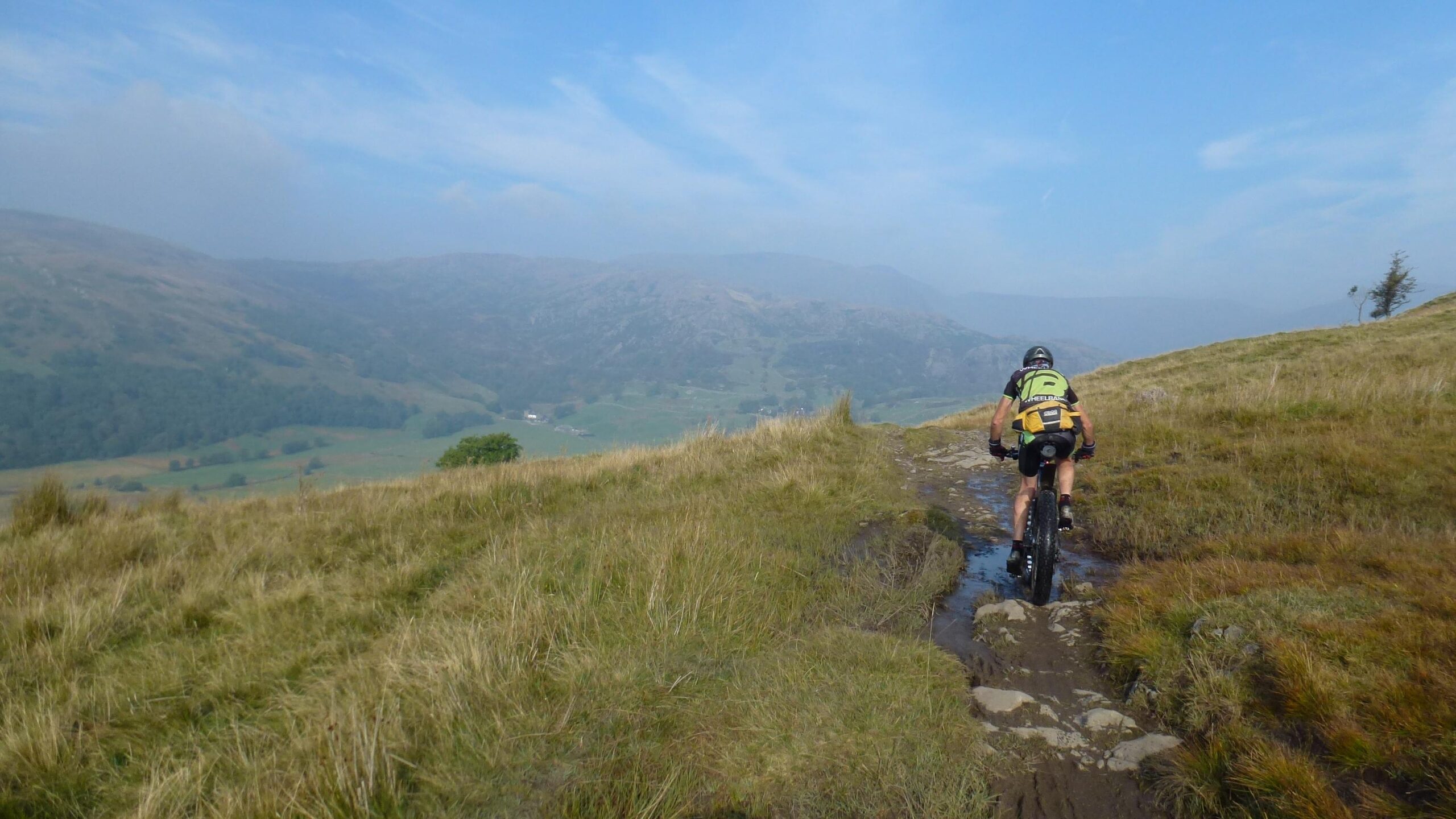 A person riding a mountain bike along a rocky path in a scenic landscape, surrounded by hills and valleys. The sky is blue with a few clouds, and the grassy terrain shows patches of mud, indicating recent rain. The rider is wearing a helmet and a backpack, enjoying an outdoor adventure in nature. Sam Houghton Circuit mountain bike trail.