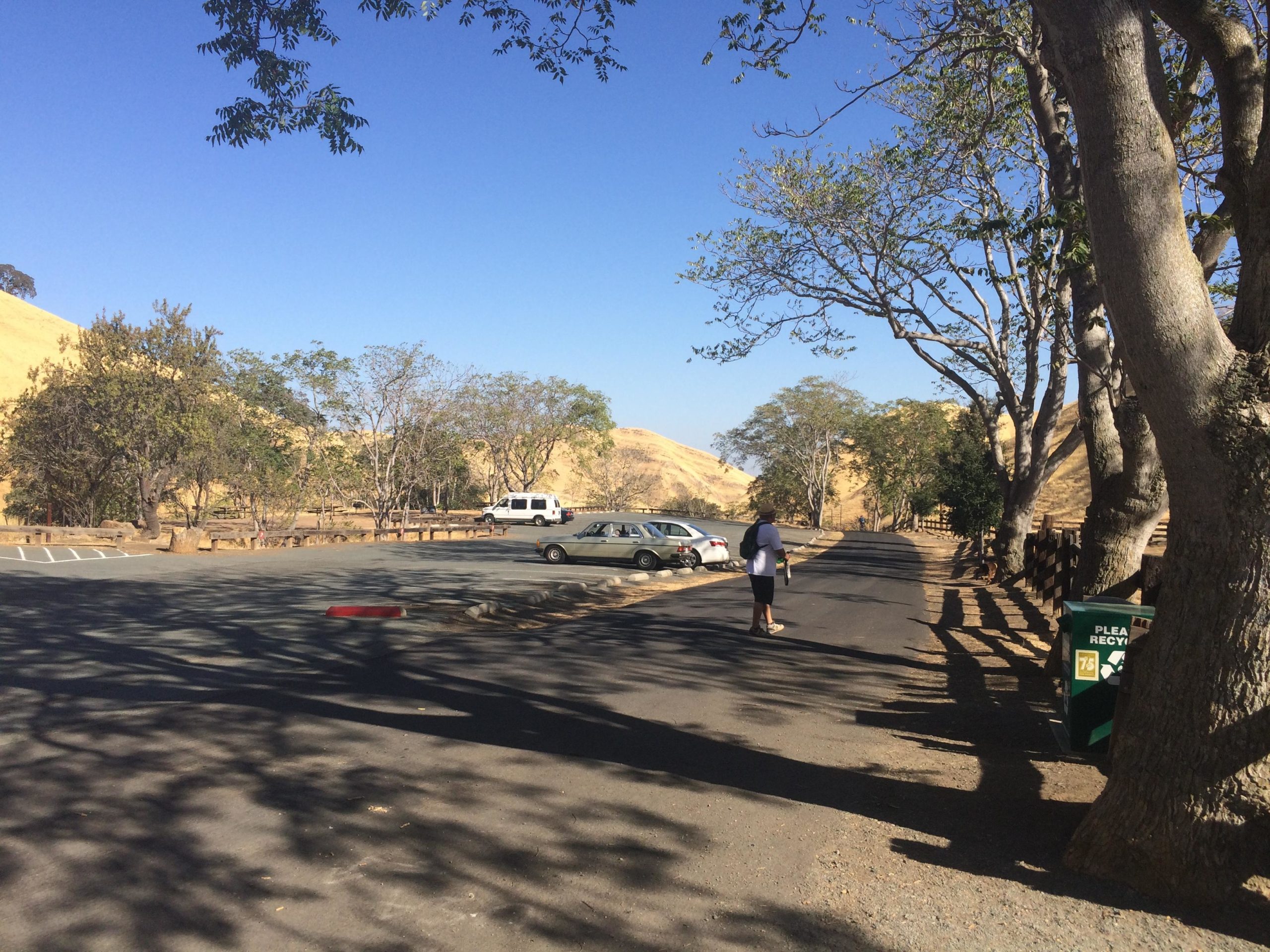 A scenic overview of a parking area flanked by trees, with a few parked cars. In the background, there are rolling hills under a clear blue sky. A person is walking along the road, and a recycling bin is visible on the right side of the image. The scene conveys a peaceful outdoor environment. Black Diamond Mines mountain bike trail.
