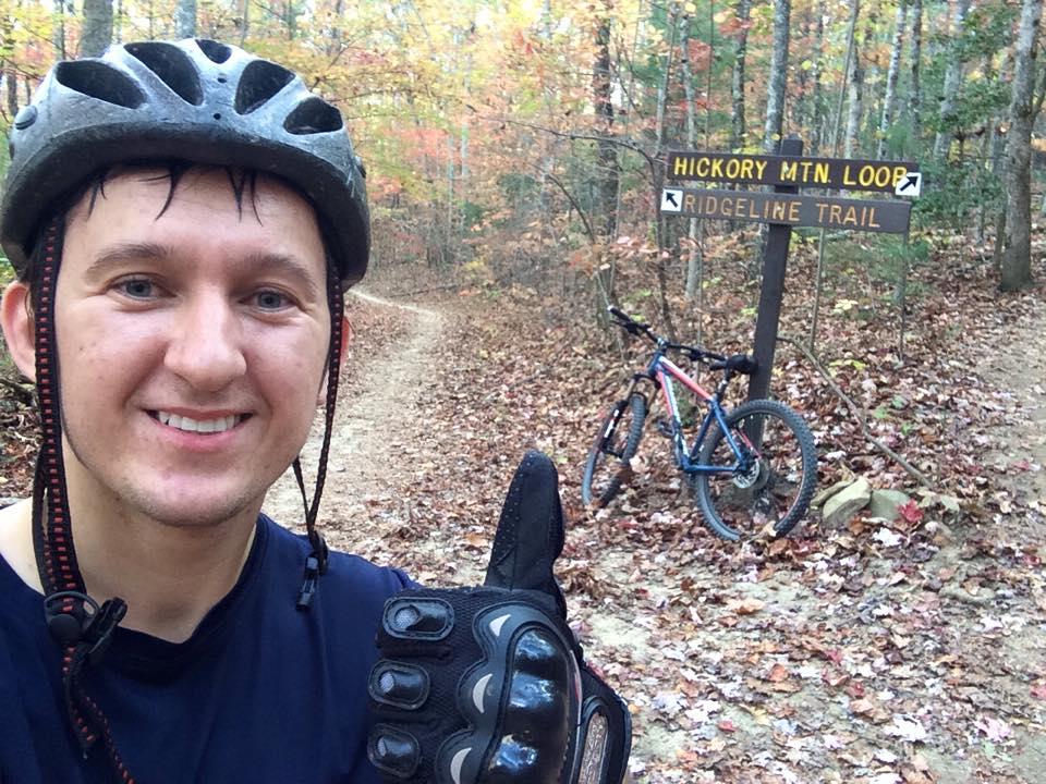 A mountain biker takes a self-portrait with a thumbs-up gesture while wearing a helmet and gloves. In the background, a wooden sign indicates the Hickory Mountain Loop and Ridgeline Trail, with a parked mountain bike nearby. The scene is set on a leaf-covered dirt path surrounded by trees displaying autumn foliage. Ridgeline Trail #65 mountain bike trail.