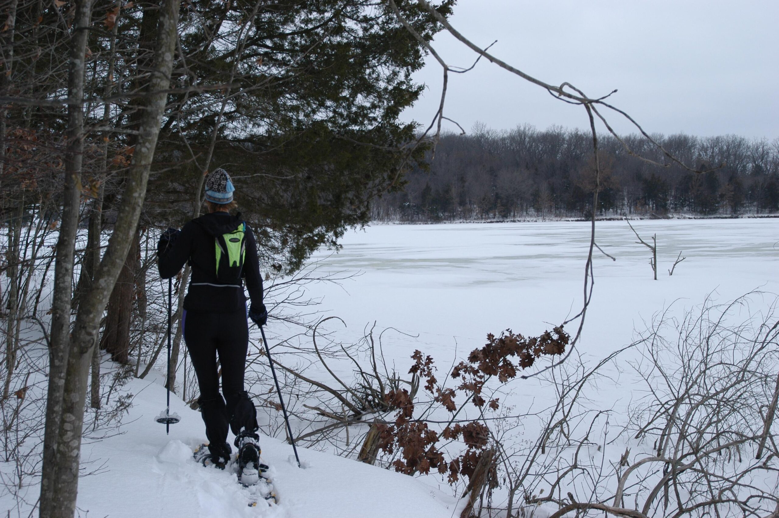A person wearing winter gear is snowshoeing near a frozen lake, surrounded by snow-covered trees and landscape. The scene is overcast, suggesting a cold winter day. Binder Lake mountain bike trail.