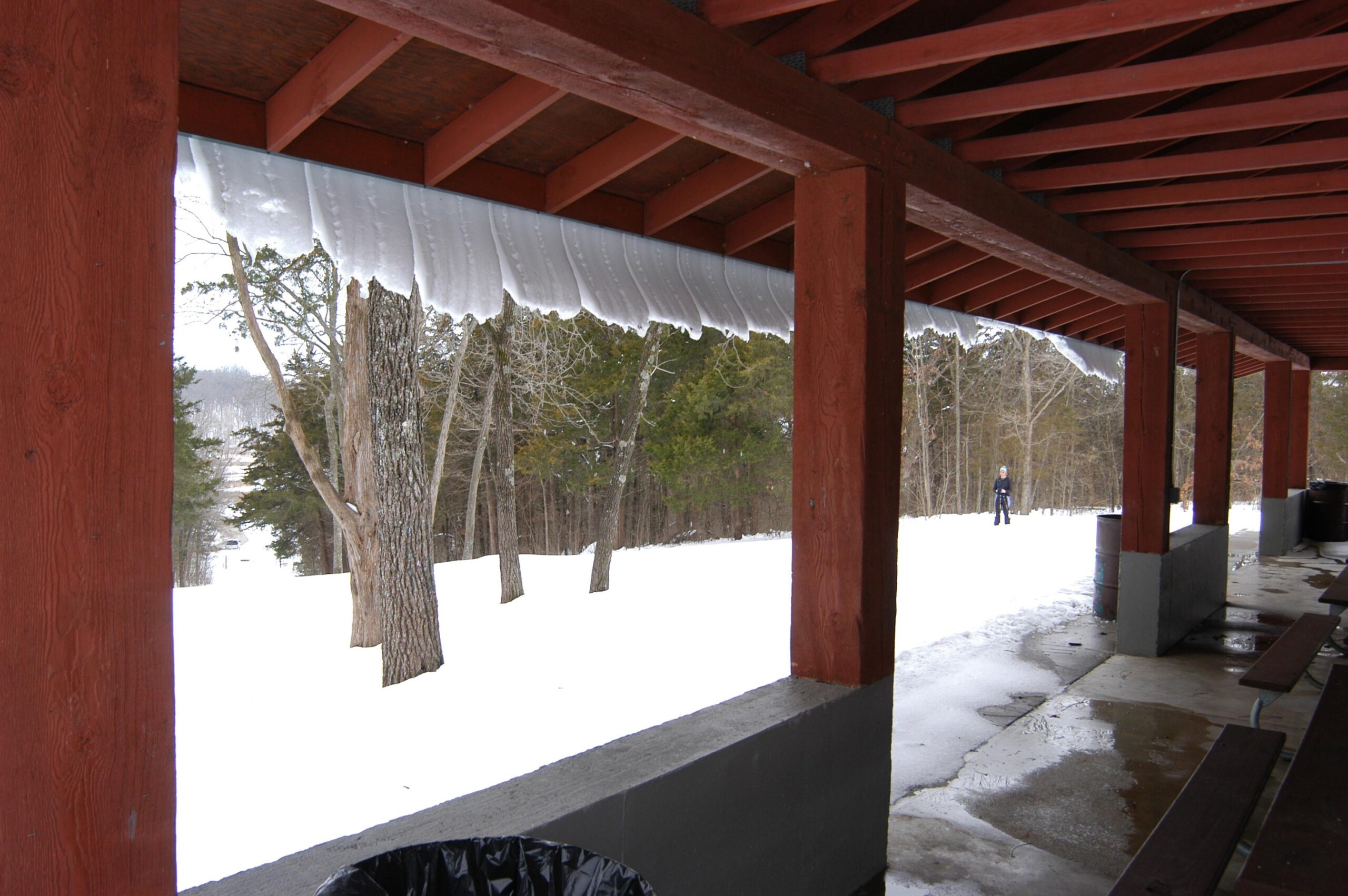 A covered outdoor area with wooden beams and a snow-covered landscape in the background. Icicles hang from the edge of the roof, and a person is seen walking in the snowy terrain. Trees are visible in the distance, and the ground shows patches of snow and wet areas. Binder Lake mountain bike trail.