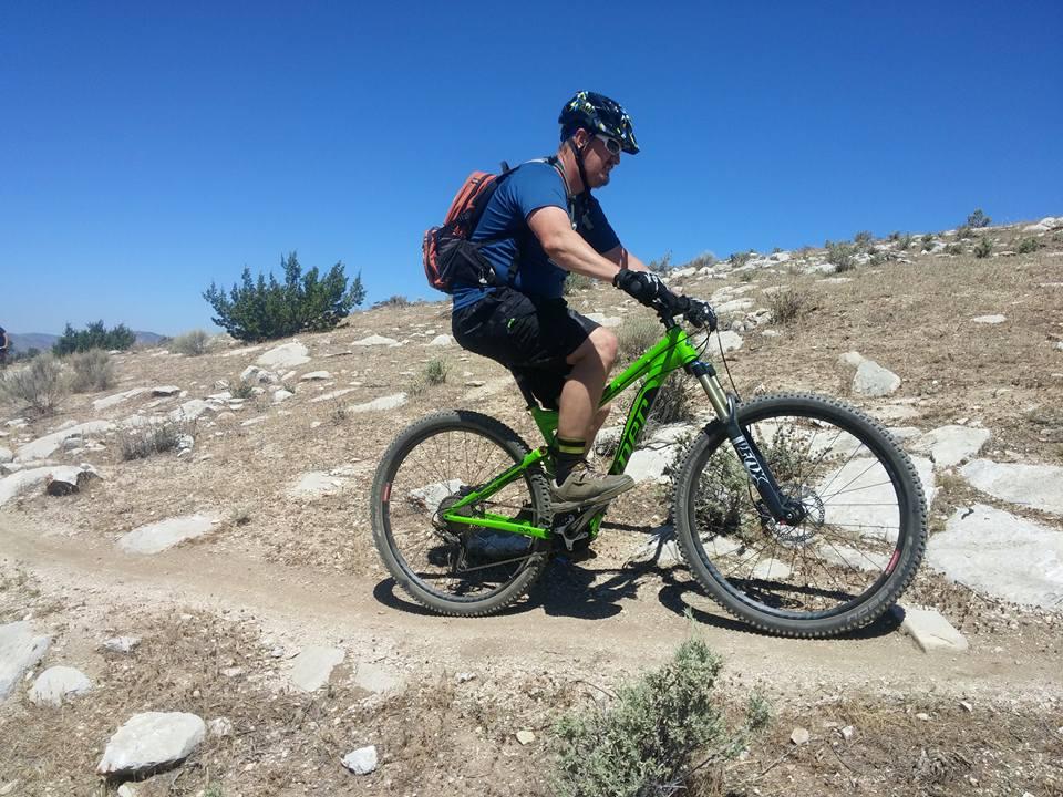 Niner RIP 9: A mountain biker riding a green bike on a dirt trail surrounded by rocks and sparse vegetation under a clear blue sky. The rider is wearing a helmet and a backpack, focusing on navigating the terrain.