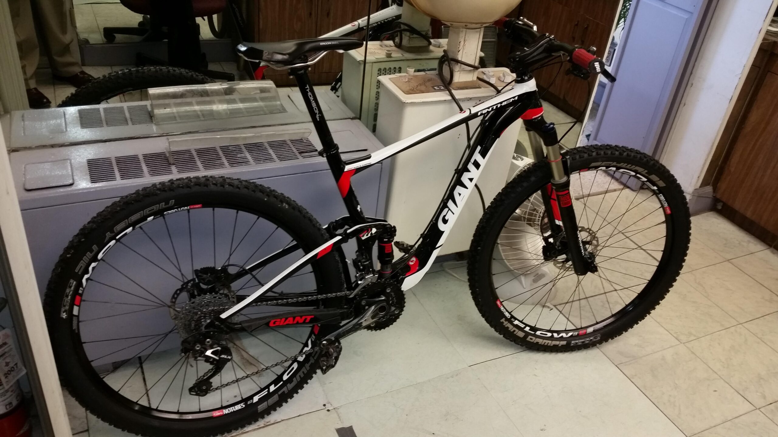 Giant Anthem: A black and white mountain bike with red accents, labeled "Giant," is positioned indoors on a tiled floor. The bike features thick, knobby tires and a suspension fork, indicating it's designed for off-road riding. In the background, various objects and furniture are visible, including a fan and what appears to be an office environment.