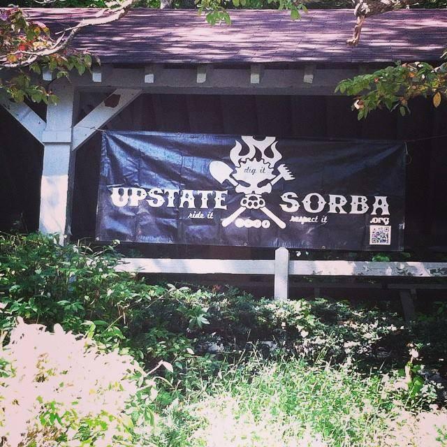 A black banner hanging from a structure, featuring the text "UPSTATE SORBA" with a graphic of a skull and crossed tools above a campfire. Below the main text, the phrases "dig it" and "respect it" are displayed. Surrounding the banner are green bushes and plants.