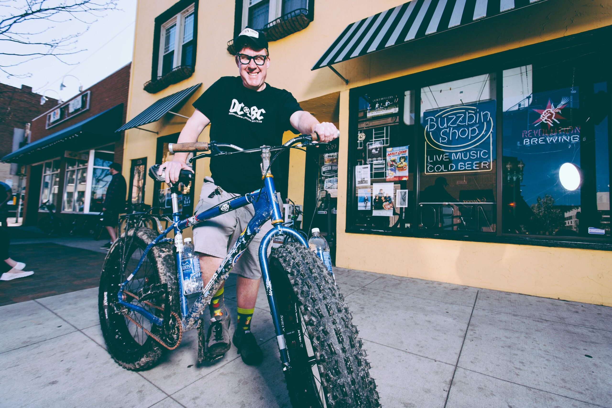 Salsa Mukluk 3: A smiling person wearing a black t-shirt and a cap stands next to a blue mountain bike with large tires, parked on the sidewalk. Behind them, there is a storefront with yellow walls and a sign that reads "Buzzbin Shop" featuring a neon sign for live music and cold beer. Two water bottles are attached to the bike, and people can be seen in the background.