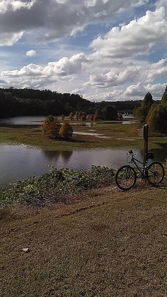 A scenic view of a tranquil lake surrounded by greenery and colorful trees, with a partly cloudy sky above. In the foreground, a bicycle rests on the grass near the water