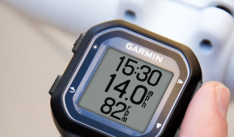 A close-up image of a Garmin watch displaying workout metrics, including time (15:30), speed (14.0 mph), and heart rate (82 bpm), with a person's hand holding the watch. The background features a blurred bicycle component.