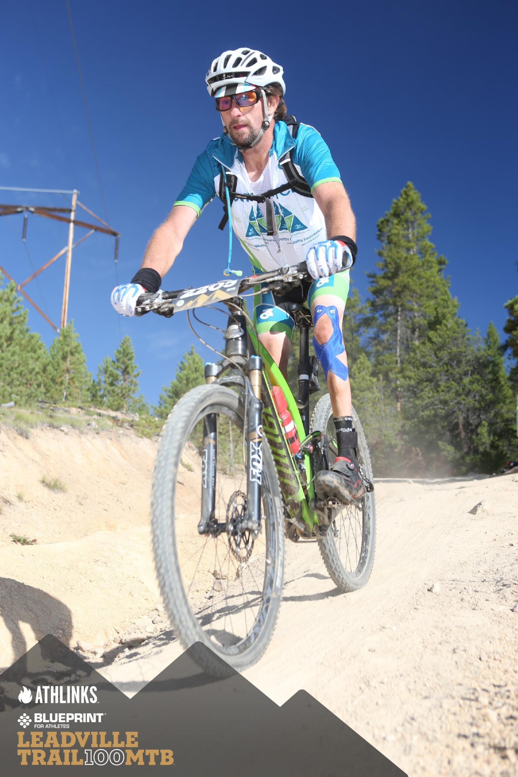 Niner Jet 9 RDO: A cyclist wearing a blue and white jersey and helmet navigates a sandy trail with trees and blue sky in the background. The cyclist's knees are wrapped in blue tape, and the bike features green and black elements. A power line structure is visible in the upper left corner of the image.