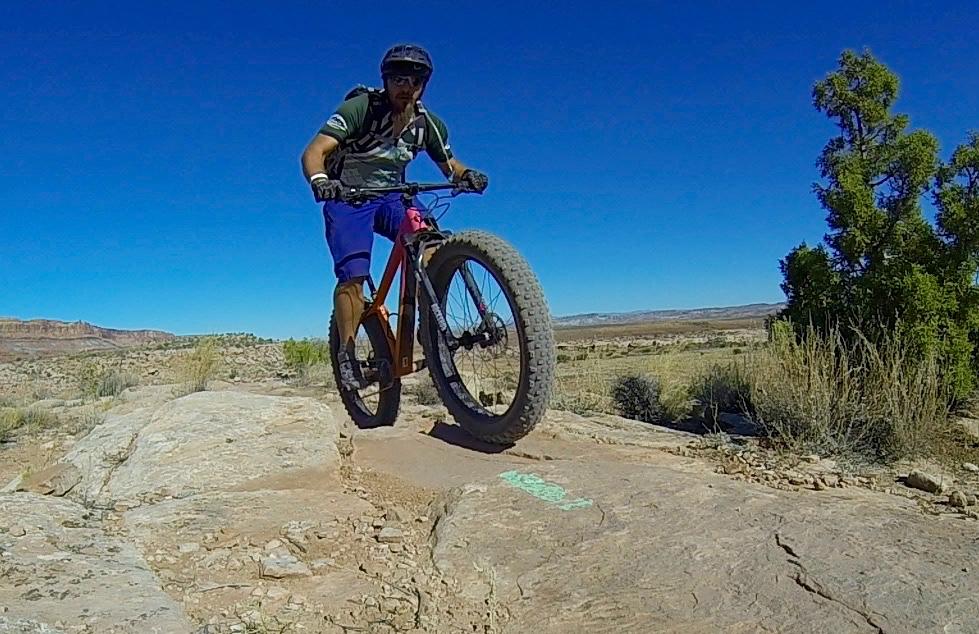 A person riding a mountain bike over a rocky terrain in a sunny outdoor setting. The cyclist is wearing a helmet and a striped shirt, with a clear blue sky in the background and desert landscape visible. Moab Brand Trails mountain bike trail.