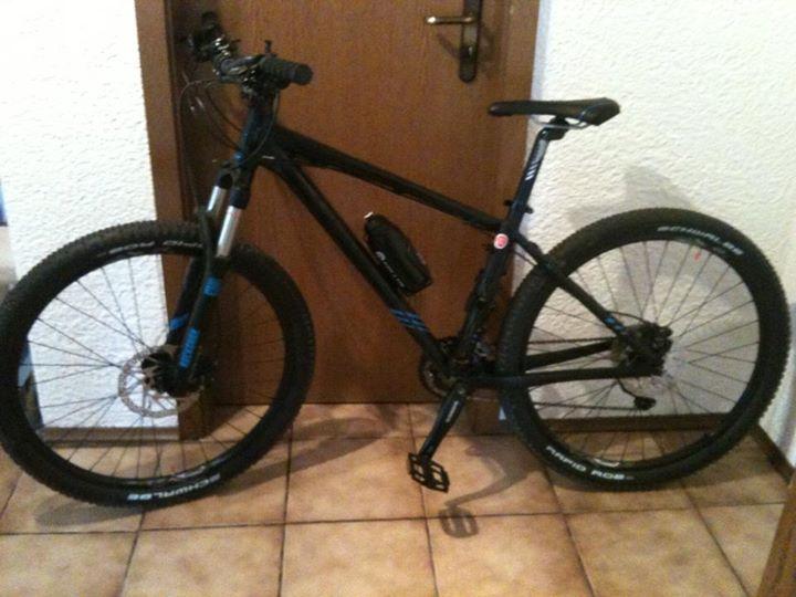Giant Talon 27.5 4: A black mountain bike with blue accents is leaning against a wooden door. The bike features thick tires, a suspension fork, and a water bottle holder attached to the frame. The background shows tiled flooring.