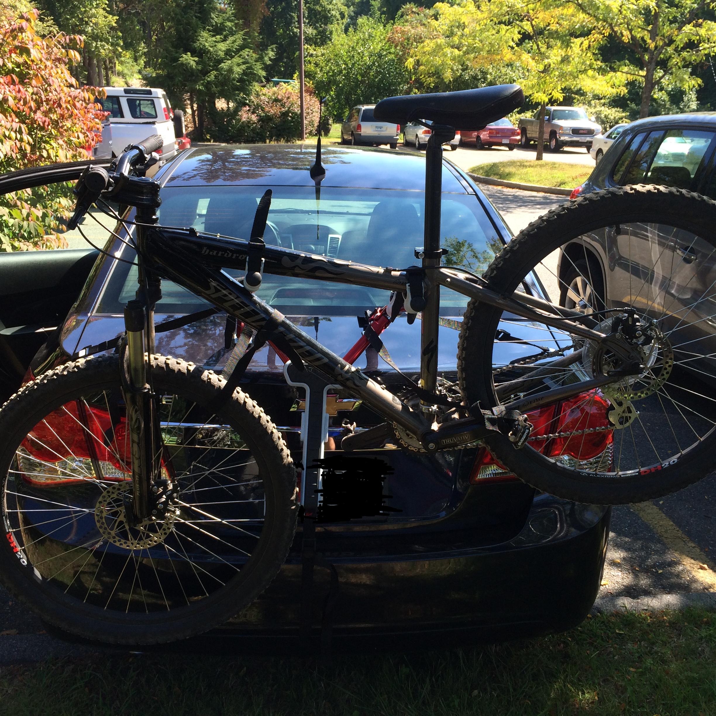 Specialized Hardrock Sport Disc: A black mountain bike secured on the back of a car, with its rear wheel and seat visible. The car's glossy surface reflects surrounding greenery and parked vehicles in a bright outdoor setting.