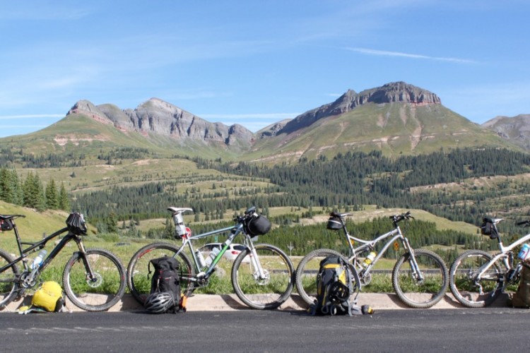 Four mountain bikes are lined up along a road with lush green hills and rugged mountains in the background. The scene is bathed in bright sunlight under a clear blue sky, with patches of trees and open fields visible in the landscape. Each bike has bags attached, suggesting they are ready for an outdoor adventure.