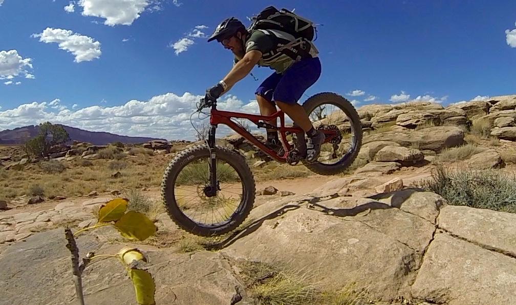 A mountain biker jumping over a rocky terrain under a blue sky filled with fluffy clouds. The rider is wearing a helmet and cycling gear, showcasing an adventurous outdoor activity in a natural landscape. Moab Brand Trails mountain bike trail.