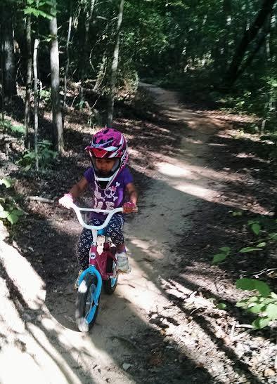 A young child riding a small bicycle on a dirt trail through a forest. The child is wearing a pink helmet and sunglasses, and is dressed in a colorful outfit. Sunlight filters through the trees, highlighting the path. North Meck Park mountain bike trail.