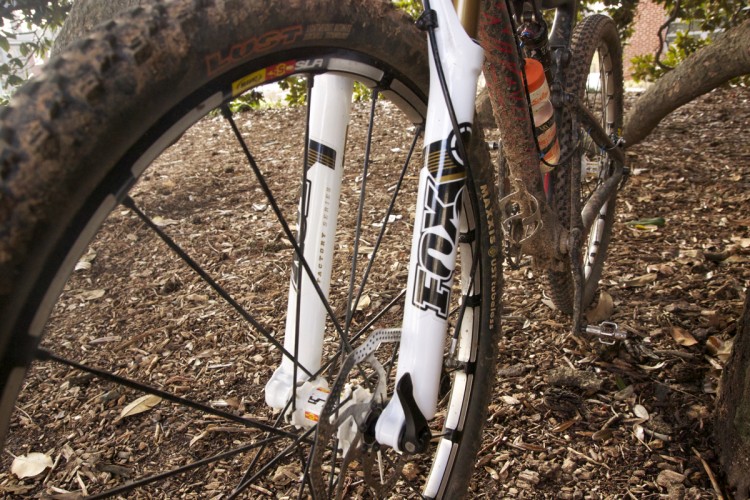 A close-up view of a mountain bike's front fork and wheel, showcasing the rugged tire and suspension components. The background features a natural setting with mulch and tree branches. The fork is branded, and the wheel has visible dirt from recent use.