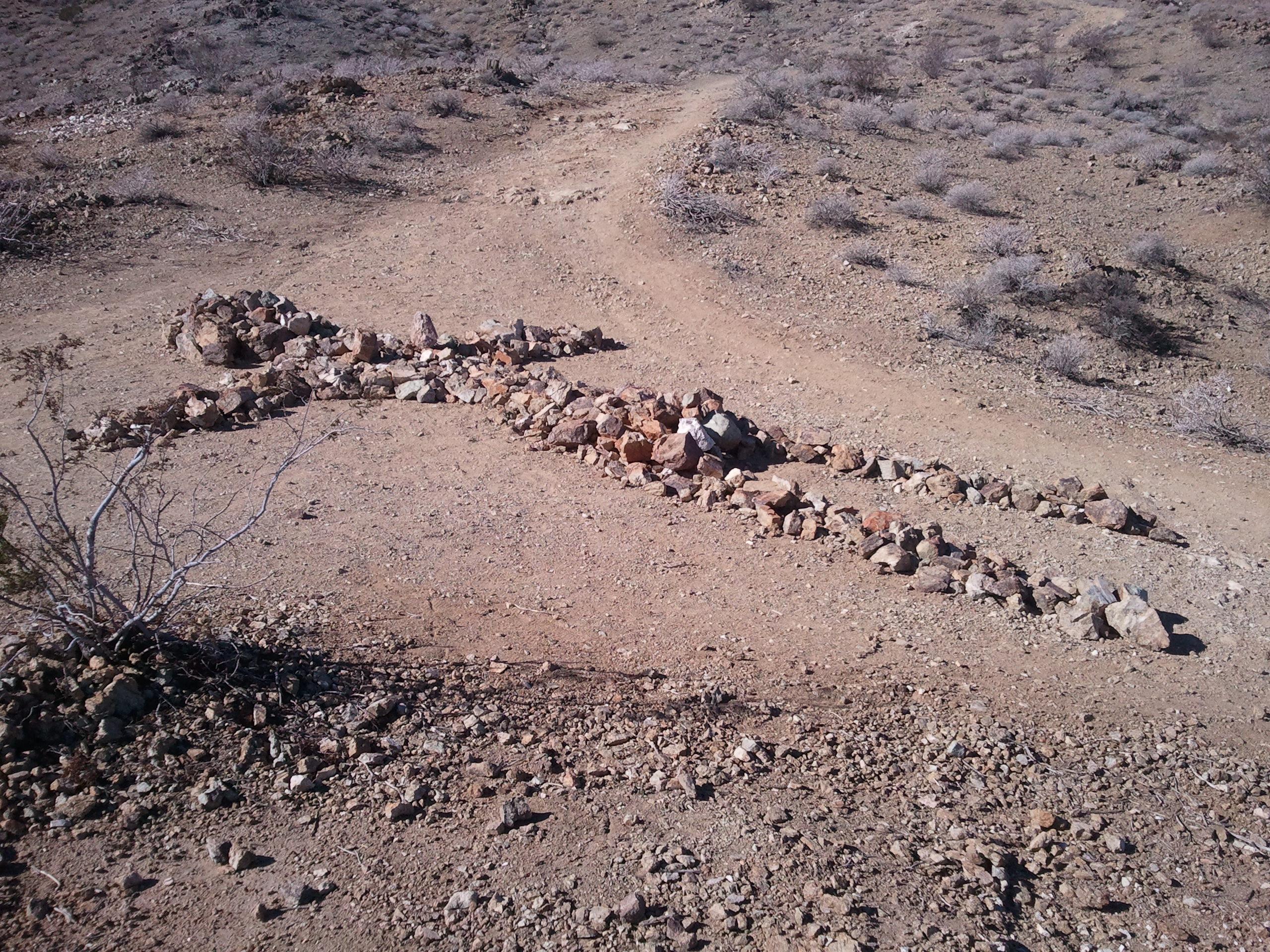 A winding arrangement of rocks on a dry, barren landscape, resembling a snake, with sparse vegetation and dirt paths in the background. The Goat Trails mountain bike trail.