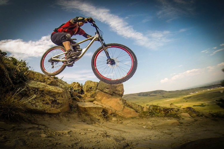 A mountain biker performing a jump over rocks, showcasing athleticism and skill against a backdrop of a blue sky and rolling hills. The rider wears protective gear and a bright red shirt, capturing a moment of action in a rugged outdoor setting.